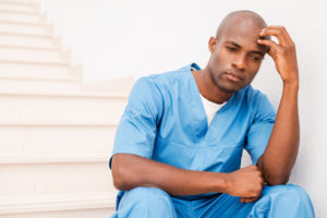 Feeling tired and depressed. Feeling tired and depressed. Depressed young African doctor in blue uniform touching his head with hand and looking away while sitting at the staircase
